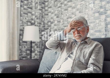 Frustrated unhealthy senior mature man touching head, having painful feelings sitting alone at home. Unhappy middle aged retired grandfather suffering from high blood pressure disease indoors. Stock Photo