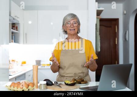 Portrait headshot of old mature woman cooking and preparing meal talking to the camera explaining her recipe to the internet online using laptop. Stock Photo