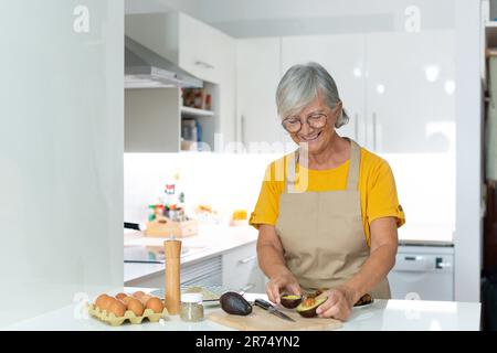 Smiling dreamy middle aged mature woman in eyewear looking in distance, distracted from chopping vegetables for salad, visualizing future or planning weekend enjoying cooking alone in modern kitchen. Stock Photo