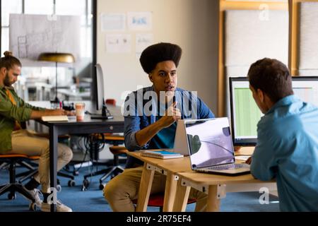 Diverse male colleagues in discussion at desks using laptops in office Stock Photo