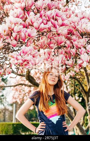 Spring portrait of adorable red-haired preteen kid girl with magnolia flowers, child wearing eyeglasses Stock Photo