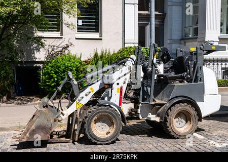 A small white excavator standing on the sidewalk near the entrance to a residential building. Stock Photo