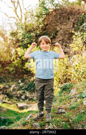 Cute little boy playing in summer park, green nature background, wearing blue t-shirt and khaki trousers, showing arm muscles Stock Photo