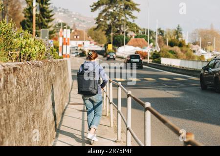 Young teenage girl riding scooter next to road, wearing backpack, back view Stock Photo