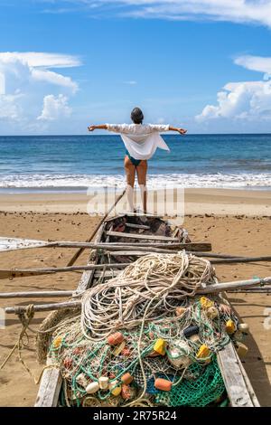 North America, Caribbean, Greater Antilles, Hispaniola Island, Dominican Republic, Sama Province, Sama Peninsula, Playa El Valle, Beautiful woman standing with arms spread on the bow of an old fishing boat looking at the sea Stock Photo