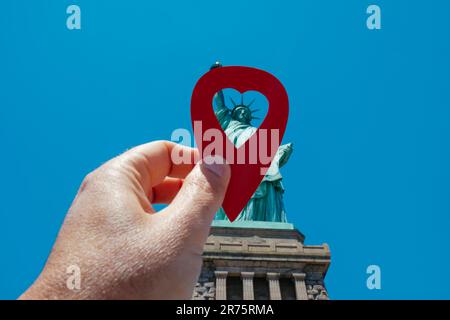 the hand of a man holding a red marker with a heart pointing the Statue of Liberty, since Liberty Island, in New York, United States, on a sunny sprin Stock Photo