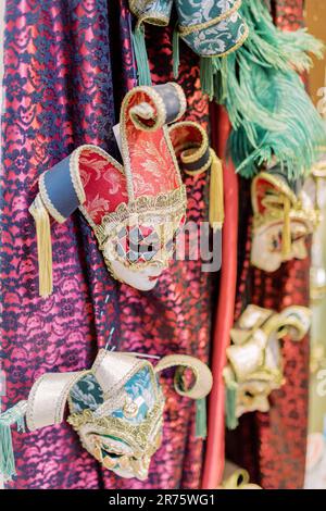 Venetian masks hang from the ceiling. Different colored fabrics can be seen in the background. The masks are colourful and ready for a masked ball. Stock Photo