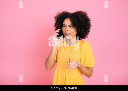 Phone talk. Confused successful busy mixed race curly haired woman, talking on mobile phone with friend or colleague, looking away, gesturing with hand, standing on isolated pink background Stock Photo