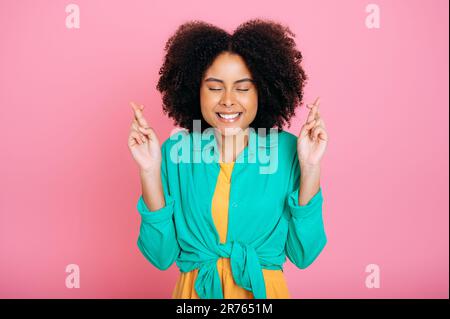 Nervous pretty brazilian or african american woman in green shirt, crossing fingers with eyes closed, praying or making wish, hope for good luck, smile, standing on pink isolated background Stock Photo