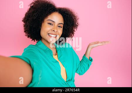 Joyful curly african american or brazilian woman, shows her hand to the empty space for your advertisement, announcement, presentation while takes selfie, stand on isolated pink background, smiles Stock Photo