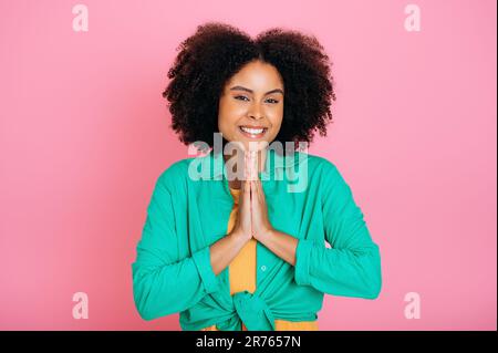 Positive pretty brazilian or african american woman in green shirt, put her palms together, praying or making wish, hope for good luck, looks at camera, smile, standing on pink isolated background Stock Photo