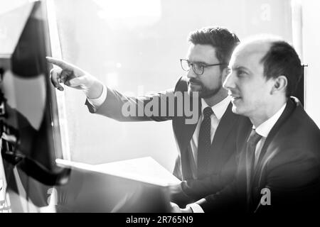 Image of two thoughtful businessmen looking at data on multiple computer screens, solving business issue at business meeting in modern corporate office. Business success concept. Stock Photo