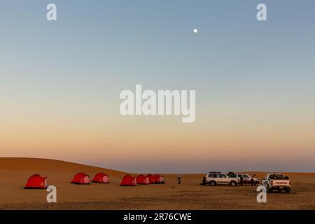 wild camping in the desert in sudan a cluster of small orange tents in the shelter of a dune with four by four vehicles under a full moon and colourfu Stock Photo