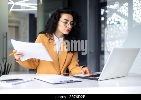 Serious focused female financier working inside office with documents and papers at workplace, hispanic woman thinking about bills on paper work. Stock Photo