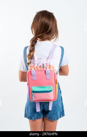 Back view of a girl in a denim suit standing with a colorful backpack on a white background in a studio Stock Photo