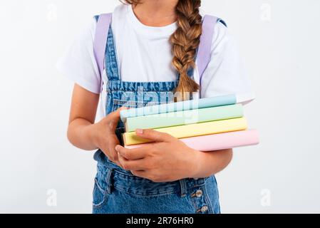 Crop unrecognizable female student in casual clothes holding books and notebook in hands while standing on white background near wall Stock Photo