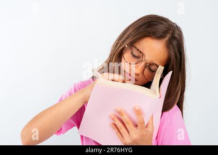 Young girl student in glasses and casual clothes reading notes in notebook while standing on white background with pen and preparing homework Stock Photo