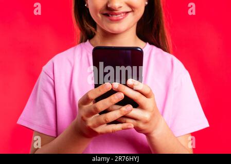 Unrecognizable cropped happy smart girl in casual outfit smiling and browsing smartphone while standing against red background Stock Photo