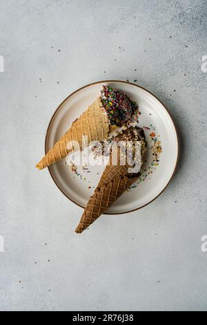 Top view of waffle cones with coffee and chocolate ice cream with multicolored sugar and nuts on a ceramic plate on a concrete background Stock Photo