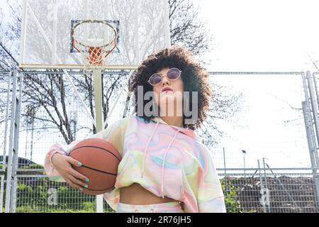 From below of young Hispanic female player in sunglasses with Afro hairstyle holding basket ball and looking at camera while standing in sports ground Stock Photo