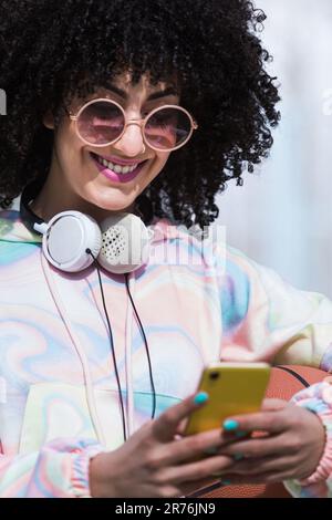 Positive young Hispanic female in Afro hairstyle sunglasses with headphones smiling and looking at screen of mobile phone while reading text messages Stock Photo