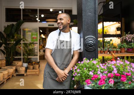 Positive young African American male florist in apron smiling and looking away while standing near blooming flowers during work day in floristry shop Stock Photo