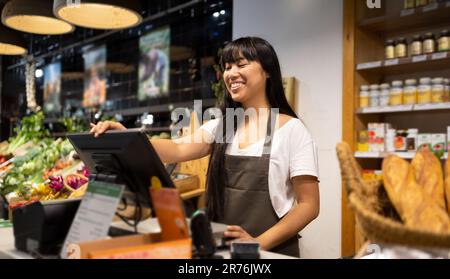 Positive young Asian female seller in apron smiling and using computer while standing at counter with various products during work in supermarket Stock Photo