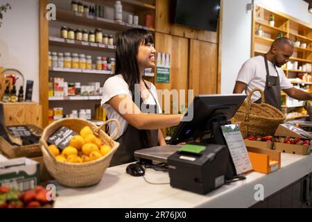 Cheerful Asian female seller in apron smiling and using computer while standing at counter with her colleague and various products while working in a Stock Photo