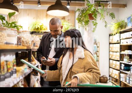 Happy young multiracial couple in casual wear smiling and choosing products while shopping together in modern store with cart Stock Photo