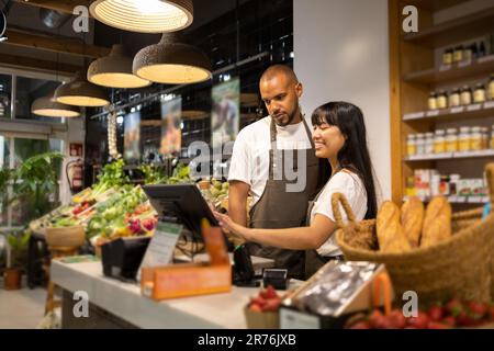 Happy young multiracial coworkers in aprons smiling and using monitor to enter information while standing at counter together during work Stock Photo