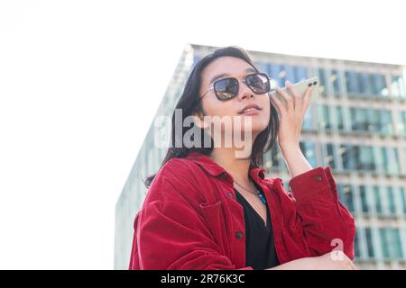 Low angle of pensive young female in casual clothes and sunglasses looking away and listening to audio message on smartphone while standing Stock Photo