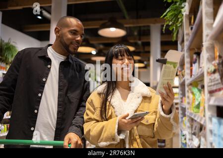 Low angle of cheerful multiethnic couple in casual clothes with shopping trolley and smartphone choosing goods while buying products together Stock Photo