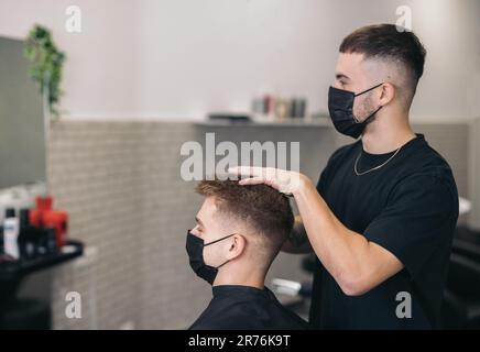 Side view of young hairdresser with mask cutting client's hair in barber shop against blurred background Stock Photo