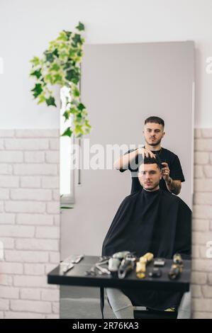Front view of reflection in mirror of young barber with tattoos combing serious client while sitting on chair in hair salon Stock Photo