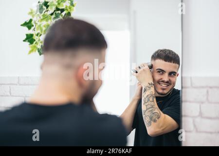 Back view of young barber cutting his hair himself while looking at mirror and smiling against blurred background Stock Photo