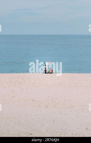 Two people enjoying a beach day, huddled together under umbrellas while looking out at the ocean Stock Photo