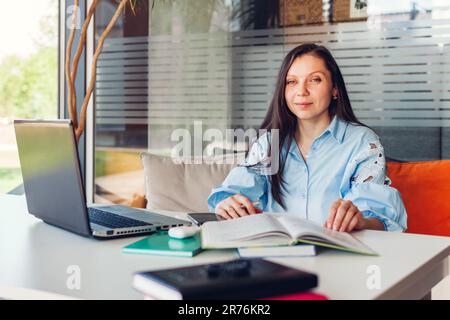 Young business woman working online in office using laptop. Middle-aged woman teacher looks at camera sitting at desk. Stock Photo
