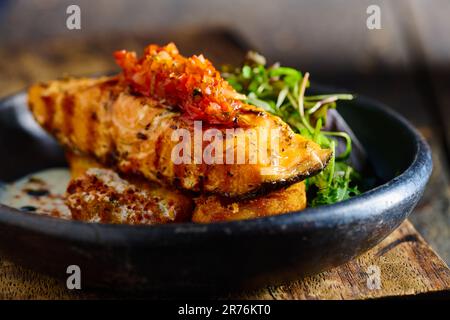 Front view of appetizing grilled salmon steak garnished with sauce and herbs served on black bowl placed on wooden cutting board Stock Photo