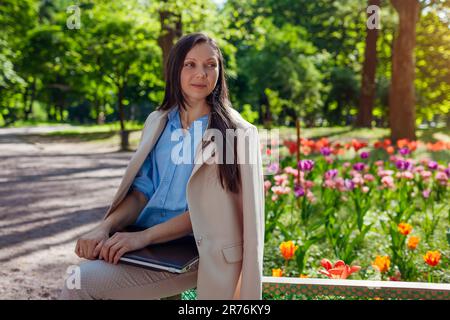 Outdoor portrait of young businesswoman working in spring park on laptop. Female landscape designer enjoys results of blooming flowerbeds in public ga Stock Photo