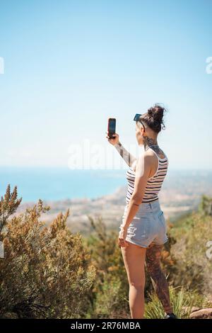 Back view of young woman in casual clothes with stylish hairdo and tattoo taking photo on smartphone while standing in countryside Stock Photo