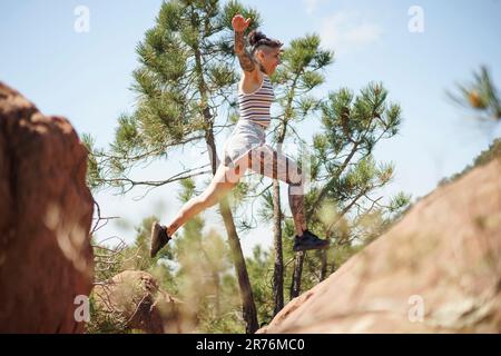 Side view of young female in casual clothes jumping over cliff high above ground with stretched arms on sunny day in countryside background with trees Stock Photo