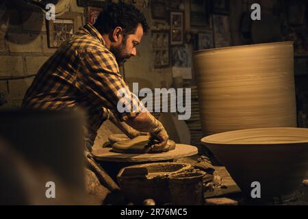 Side view concentrated of middle aged ethnic bearded male master in checkered shirt and apron molding clay while creating pots in traditional pottery Stock Photo