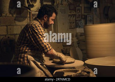 Side view concentrated middle aged ethnic bearded male master in checkered shirt and apron molding clay while creating pots in traditional pottery Stock Photo