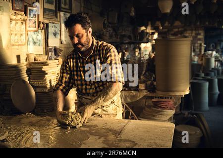 Concentrated middle aged ethnic bearded male master in checkered shirt and apron molding clay while creating pots in traditional pottery Stock Photo