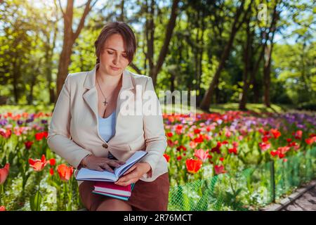 Young caucasian business woman ckecking notes in spring park. Teacher having break outdoors. Landscape designer working outdoors Stock Photo