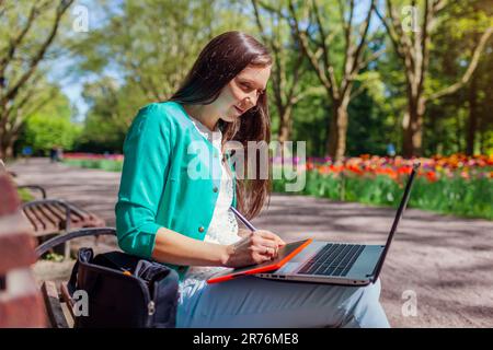 Outdoor portrait of student studying in spring park using laptop and tablet. Young woman getting ready for lecture in public garden Stock Photo