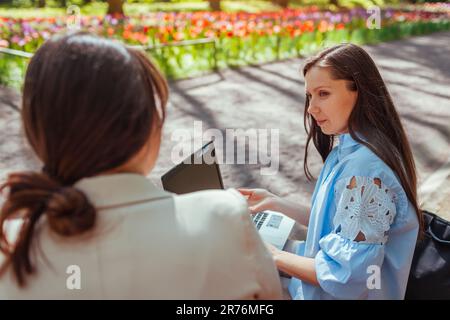 Two young businesswomen cowokers working in spring park using laptop and book. Remote work during break. Friends discussing tasks. Team partner meetin Stock Photo