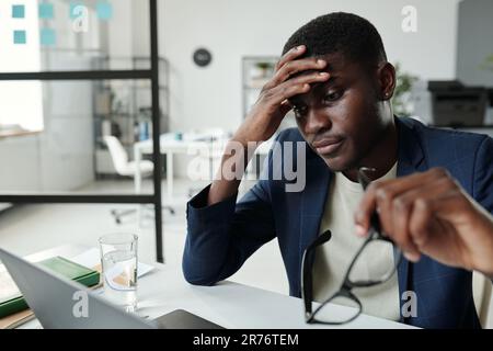 Young tired businessman or student touching forehead while sitting by desk in front of laptop and looking through online data on screen Stock Photo