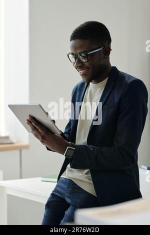 Young smiling male manager in eyeglasses and formalwear using tablet while looking through online information and analyzing it in office Stock Photo