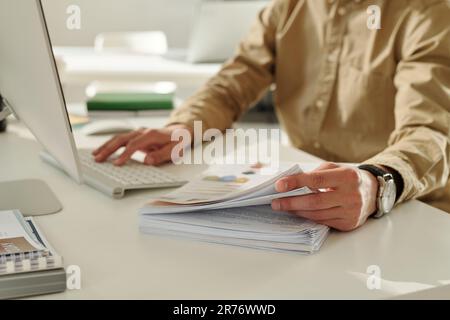 Focus on hand of young financial director of marketing company taking some documents from stack of papers while working by desk Stock Photo
